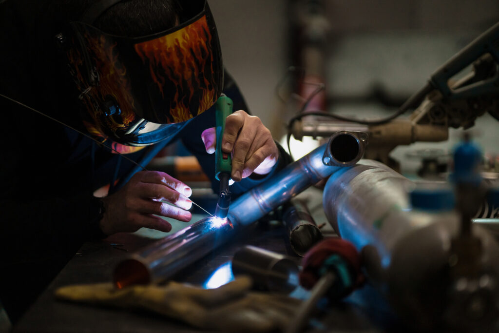 Worker in helmet with flames welding the steel part of a motorbike using an argon welding machine and gloves in a garage